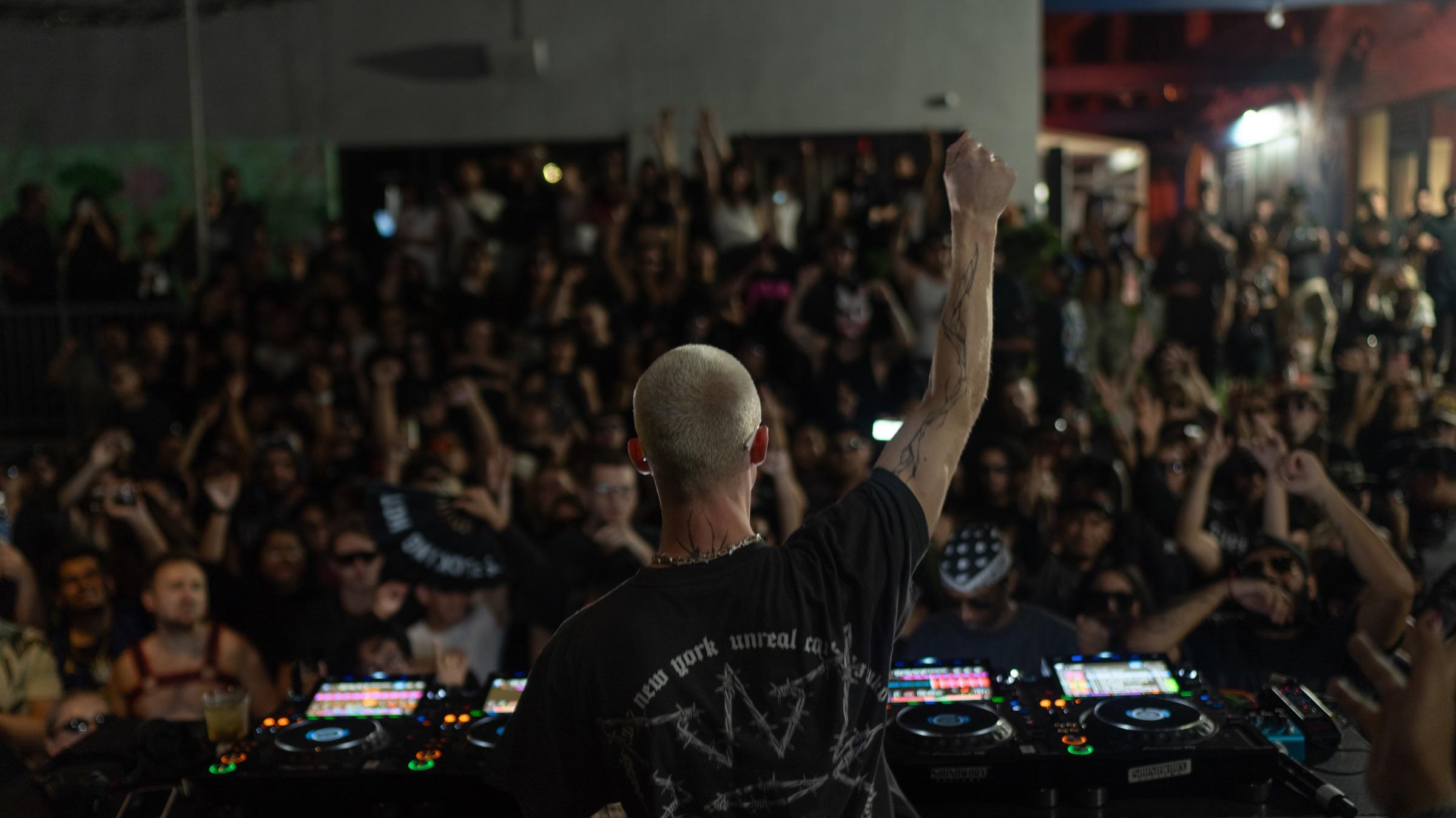 Crowd seen from behind the booth as the DJ raises an arm