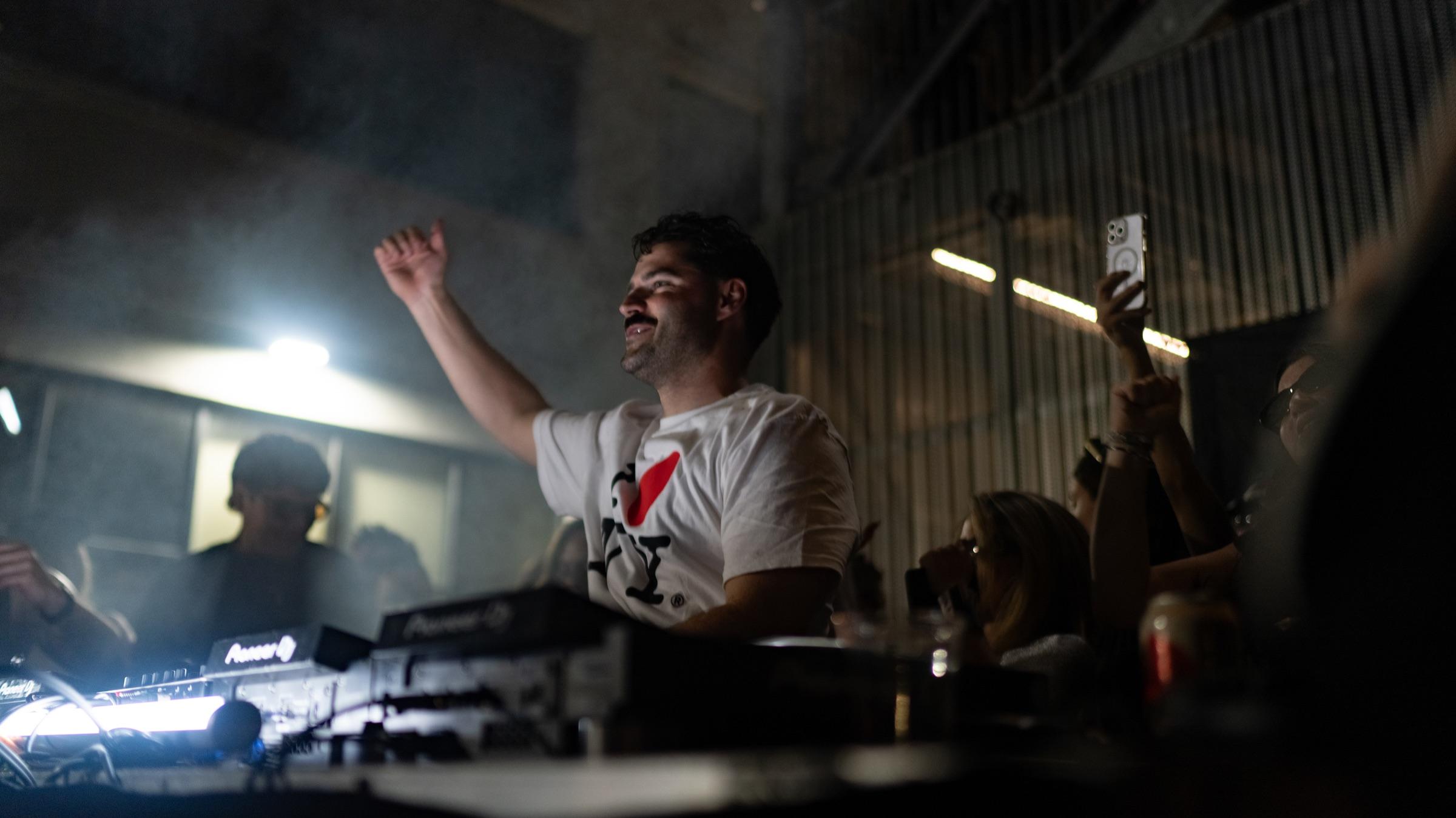 DJ with arm raised at the booth, crowd behind