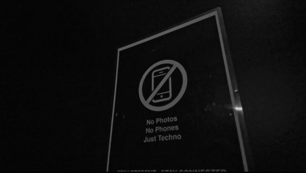 Black-and-white sign in the dark: No photos, no phones, just techno