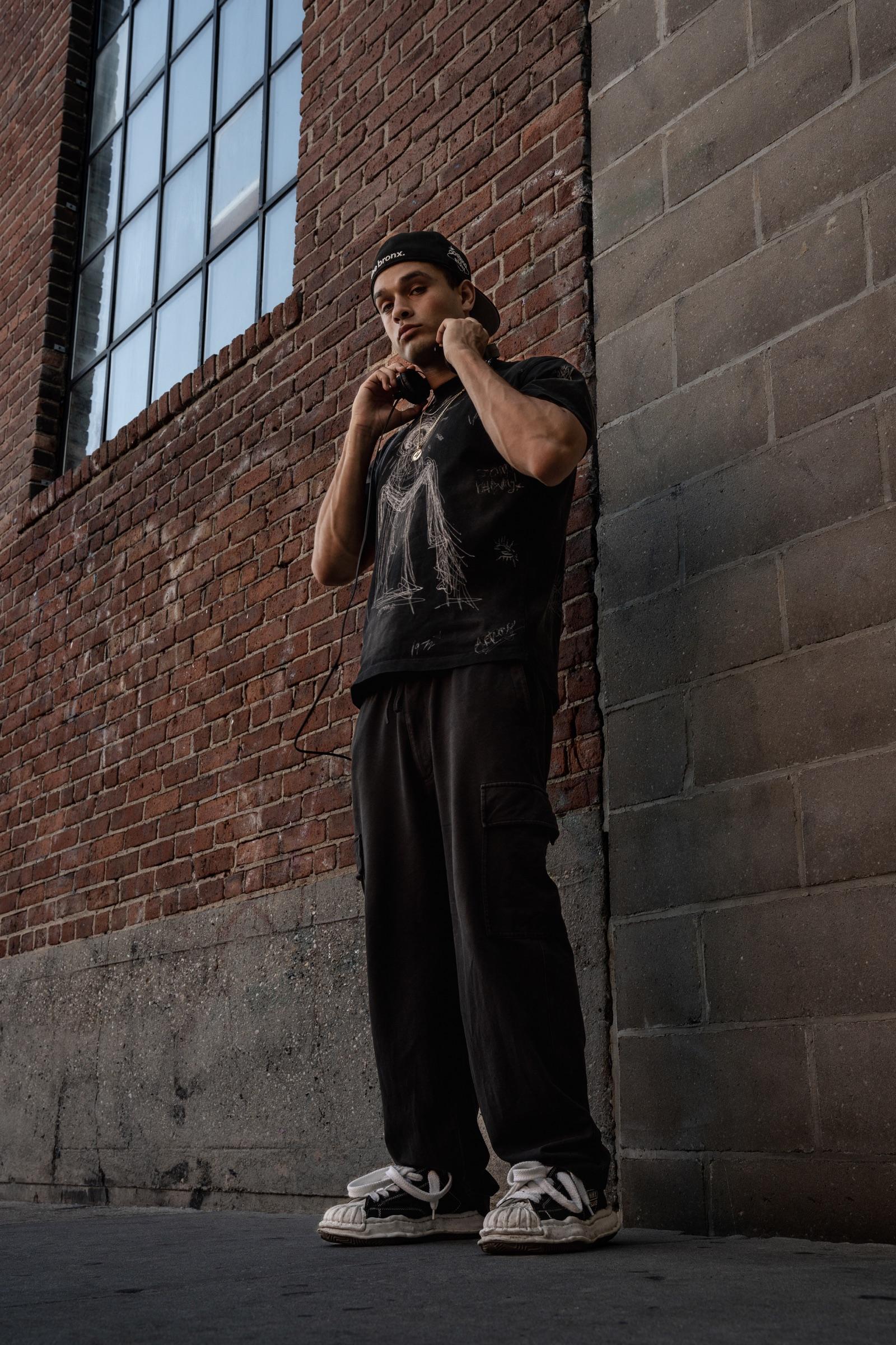 Artist standing against a brick wall, hands on headphones