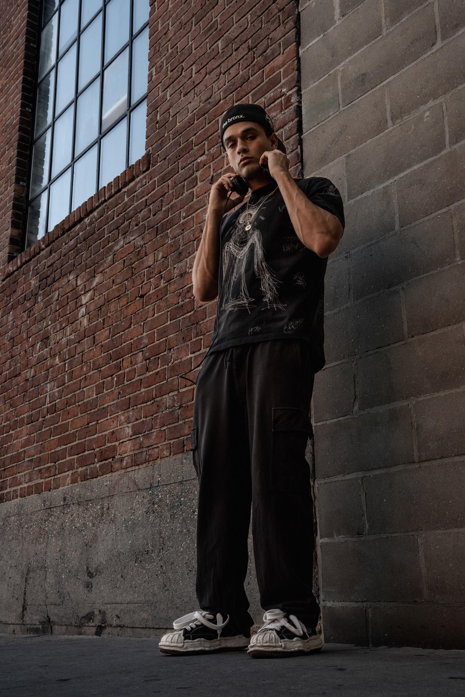 Artist standing against a brick wall, hands on headphones
