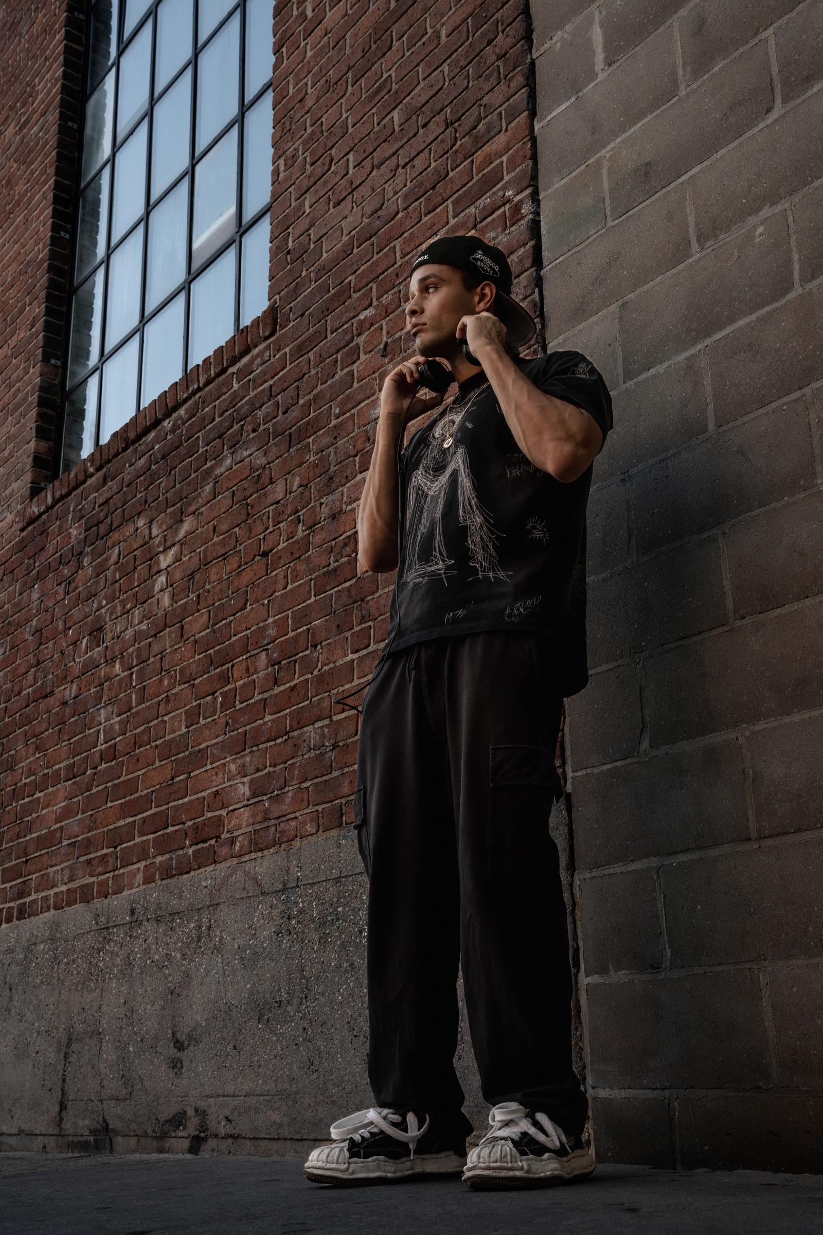 Artist standing against a brick wall, hands on headphones