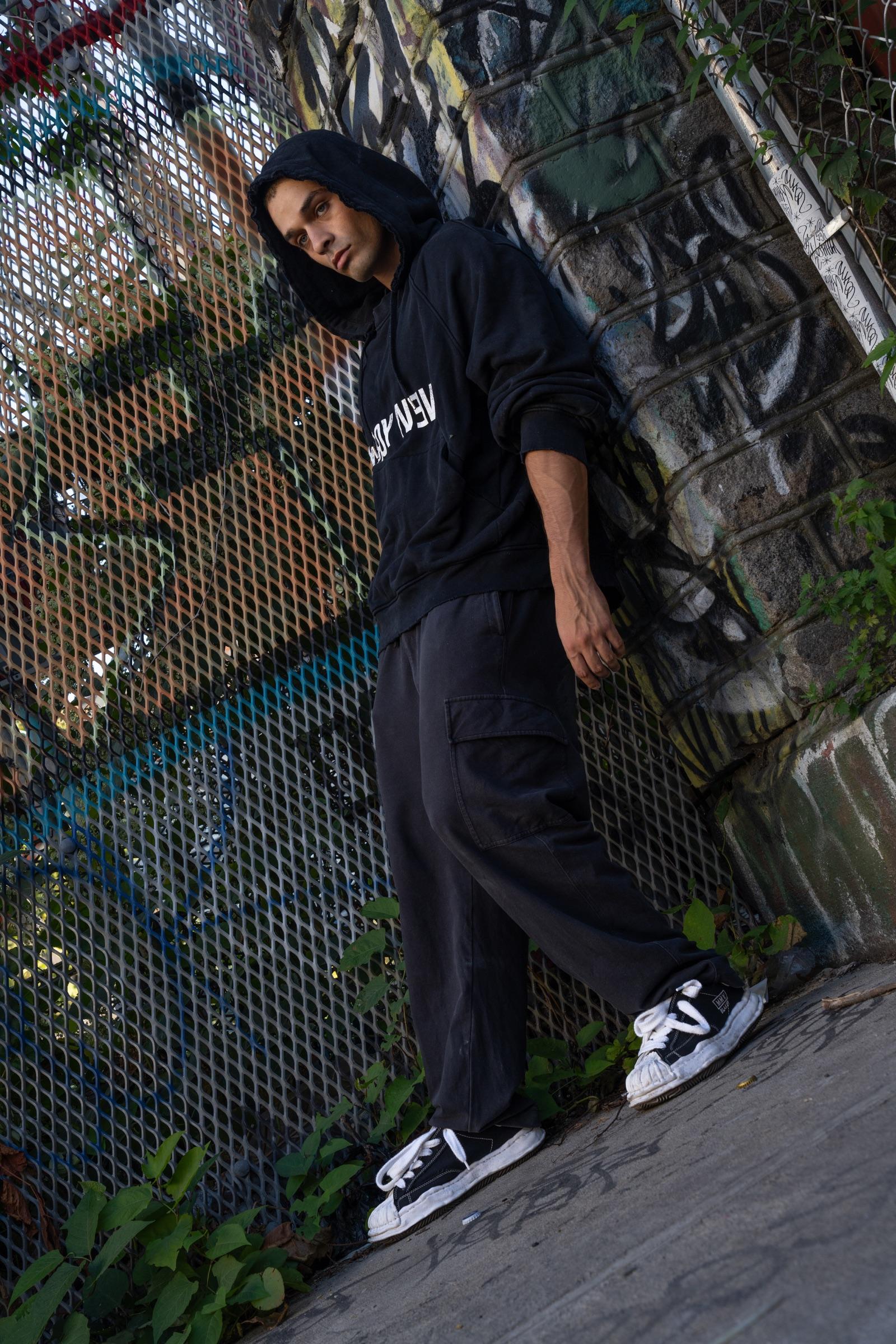 Artist in a hood leaning against a graffiti fence