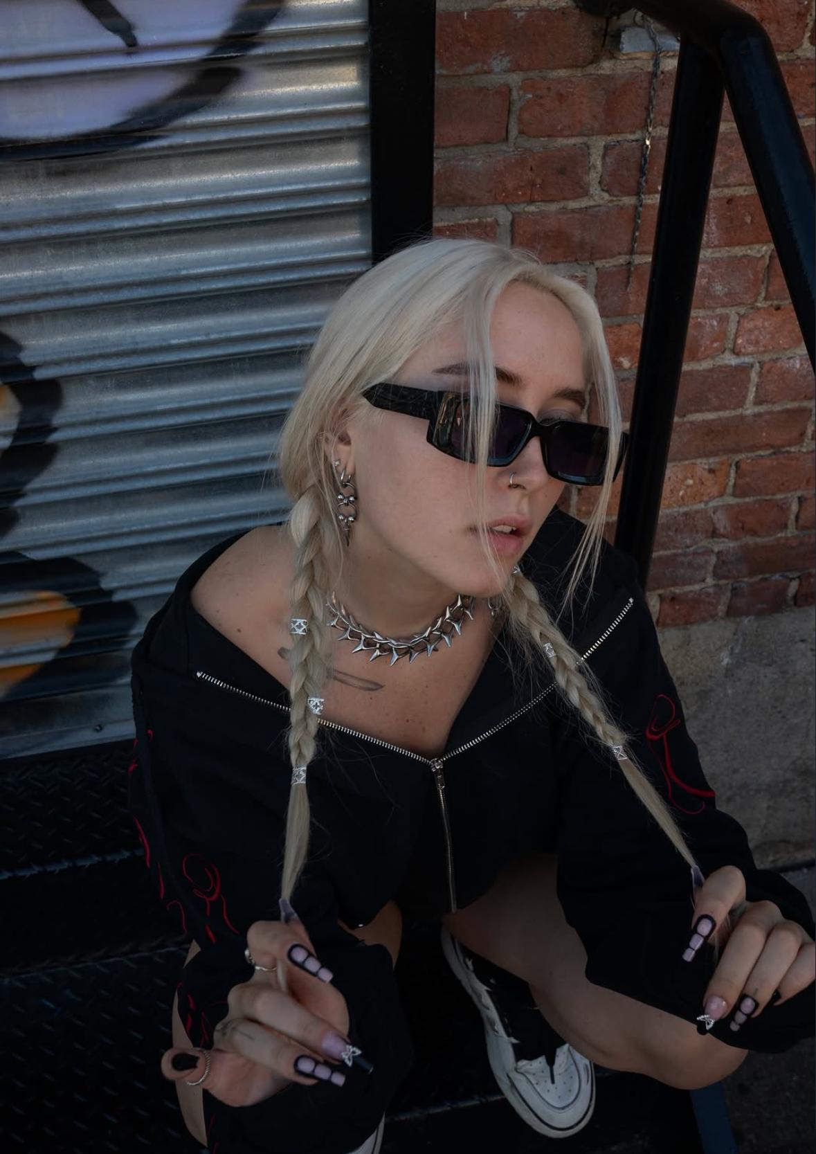 Portrait of Marilyn in sunglasses with blonde braids, sitting on steps by a brick wall
